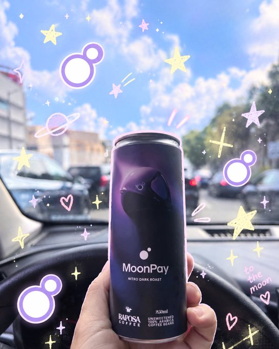 A hand holding a 230ml can of MoonPay Nitro Dark Roast by Raposa Coffee against a car dashboard background, decorated with playful purple and yellow doodles and 'to the moon' text.