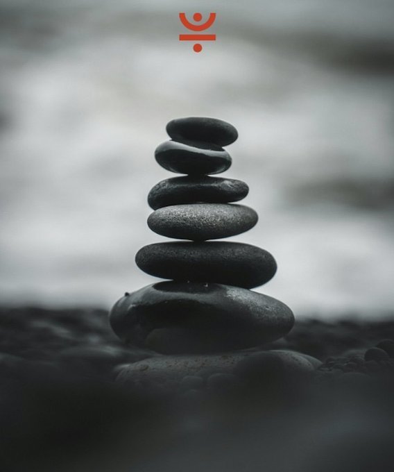 A serene image of a stack of smooth black stones balanced on a beach with the ocean in the blurred background, featuring a small red logo at the top.