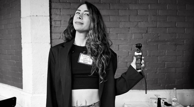 A black-and-white photo features a woman with long wavy hair wearing a black blazer over a "Burger Records" t-shirt, holding a small camera.