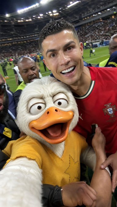 Cristiano Ronaldo smiling and posing for a selfie with a duck mascot wearing a yellow sweater at a stadium event.