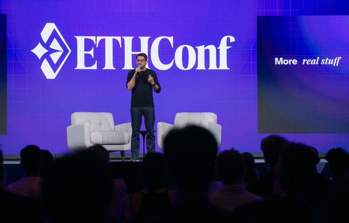 The image shows a man standing on stage at an event, giving a speech or presentation. The stage has a large screen behind him displaying the name "ETHConf" written in white letters. There are two chairs visible near the front of the stage, one closer to the camera and another further back. A crowd of people is present in the audience, attentively watching the speaker.