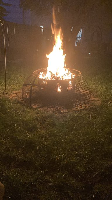 A fire pit burning brightly with tall flames on a grassy lawn at night, surrounded by a metal dome grate.