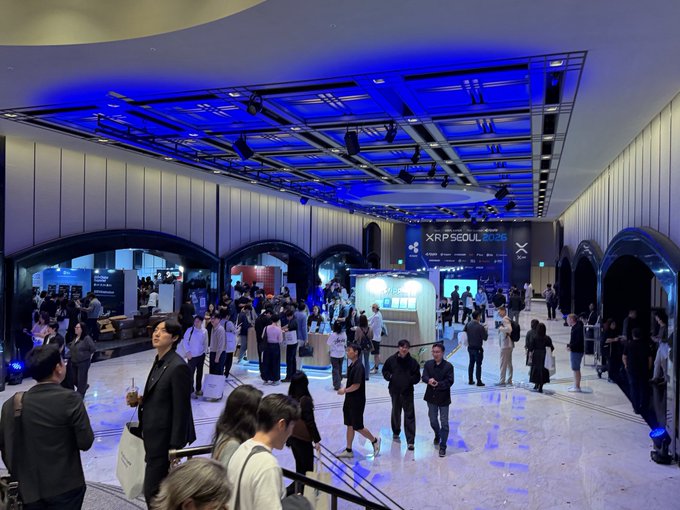 A wide shot of the XRP Seoul 2025 conference, showing a large crowded hall with blue-lit ceiling panels, exhibition booths, and attendees networking.