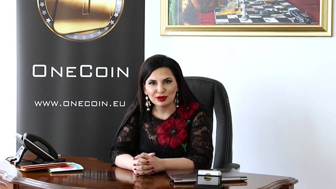 Dr. Ruja Ignatova, known as the "Crypto Queen," poses behind her desk in front of a OneCoin banner displaying "WWW.ONECOIN.EU," representing the fraudulent cryptocurrency scheme she founded that defrauded investors of $4.5 billion before her 2017 disappearance.