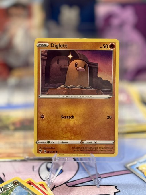 A Pokémon trading card featuring Diglett (HP 50) with a 'Scratch' attack, displayed on a clear stand with other cards blurred in the background.