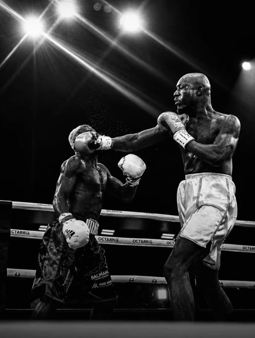 A dramatic black-and-white photograph of a boxing match in progress, showing one boxer landing a powerful punch on his opponent inside a ring with CTARRAIA and OCTABRIS branding on the ropes, illuminated by bright overhead arena lights.