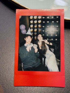 A red Polaroid-style photo of a couple wearing party hats blowing party horns, with a white dog joining them.