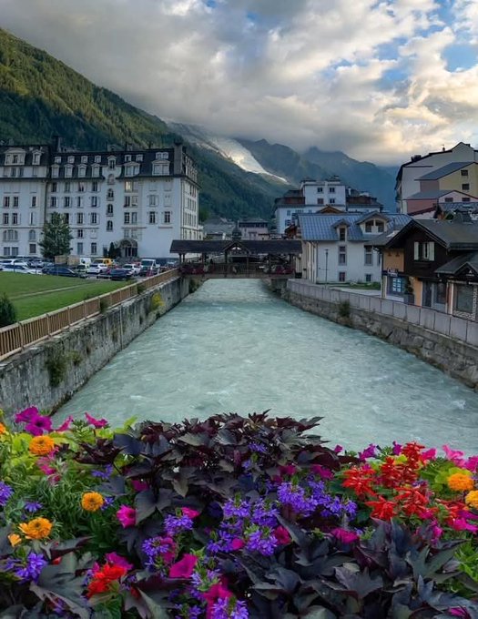 The image shows a beautiful view of a river surrounded by colorful flowers and buildings with balconies overlooking the water. In the background, there are mountains covered in clouds, adding to the picturesque scenery. The photo appears to be taken from a bridge or a similar elevated position, providing an interesting perspective on the landscape below.