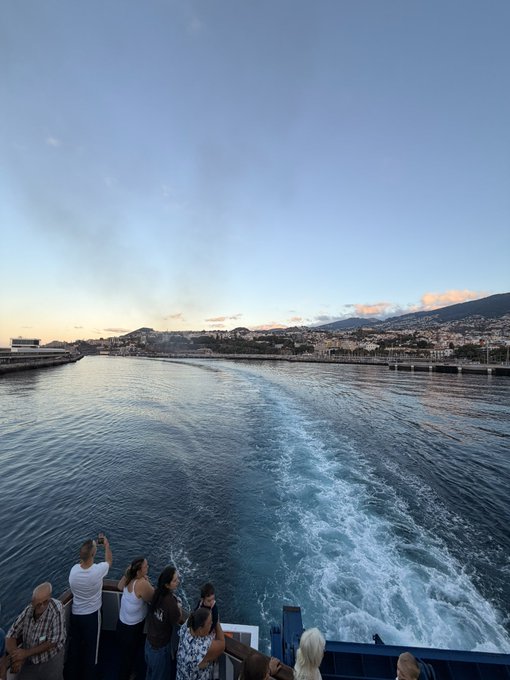 A group of passengers stand at the stern of a ferry, watching the foamy wake trail behind the vessel as it departs a coastal city bathed in golden morning light. The scenic harbor view captures the essence of a perfect 120-minute getaway, with the rugged coastline and distant mountains serving as an idyllic backdrop for the journey.