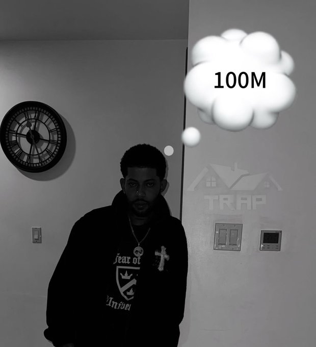 A black-and-white photo of a young man wearing a 'Fear of God' jacket and cross necklace, standing indoors near a wall clock, with a thought bubble showing '100M' and an 'AIR TRAP' watermark.