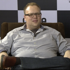 A bespectacled man in a light blue button-down shirt sits relaxed on a brown leather couch during what appears to be an interview setting. The image accompanies a news story about Japheth Dillman, who was convicted of wire fraud related to a crypto trading fund that defrauded over 20 people of nearly $1 million and now faces up to 20 years in prison and a $250,000 fine.