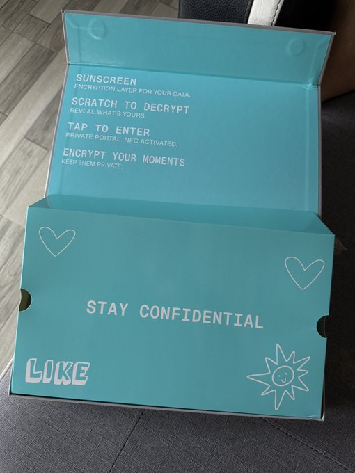 An open teal-colored box with a 'LIKE' branding, featuring instructions like 'SUNSCREEN - ENCRYPTION LAYER FOR YOUR DATA,' 'SCRATCH TO DECRYPT,' 'TAP TO ENTER,' and 'ENCRYPT YOUR MOMENTS,' along with 'STAY CONFIDENTIAL' and decorative hearts and a sun.