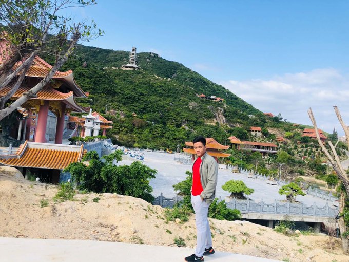 A young man in a red shirt and beige jacket posing in front of a Chinese-style temple complex with traditional orange-roofed pavilions, set against a lush green hillside.