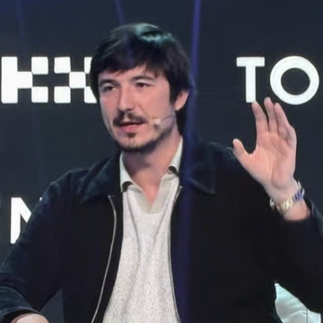 A man with dark hair and a beard, wearing a black jacket over a white shirt, speaks on stage while gesturing with his hand raised, with partial text "HO" and "TO" visible in the background.