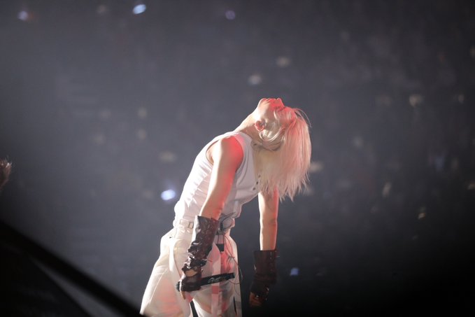 A performer with platinum blonde hair, wearing white attire and arm gauntlets, arches their back in a dramatic pose on stage. This image evokes the "AURA" surrounding a captivating live performance.