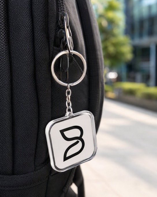 A square metallic keychain with a stylized 'B' logo attached to a black leather bag.