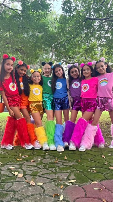 A group of eight young girls dressed in rainbow-colored Teletubbies costumes with colorful furry leg warmers, posing together outdoors.