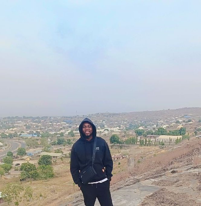 A man wearing a black hoodie, glasses, and a crossbody bag stands outdoors with a sprawling town landscape behind him.