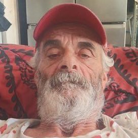 An elderly bearded man wearing a red cap reclines on a patterned red couch, his weathered face giving a subtle, knowing look to the camera. The image pairs with the caption "take a risk white boy," lending a humorous, self-deprecating tone as if the seasoned old-timer is daring the viewer to embrace boldness despite uncertainty.