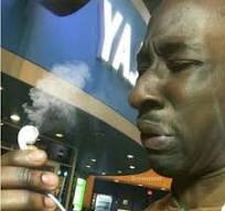 A man smoking a cigarette or blunt with a 'YAMAHA' sign visible in the background, exhaling a cloud of smoke.