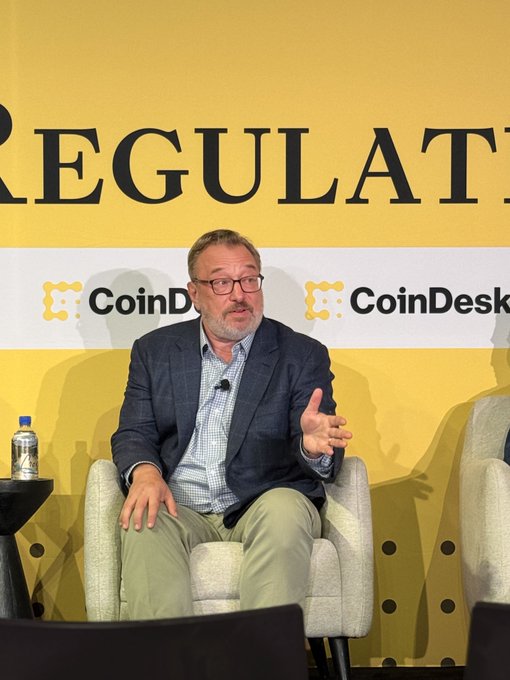 A man with glasses and a beard speaks while seated on a chair during a CoinDesk 'Regulation' panel discussion, gesturing with his right hand.