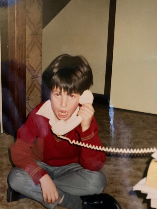 A vintage photograph shows a young boy with dark hair, sitting cross-legged on a carpeted floor while holding a white landline telephone receiver to his ear with a serious expression. The caption "This is who runs this account btw" suggests the photo is meant to portray the account's creator as a young child, humorously presenting a childhood image as a representation of who manages the account.