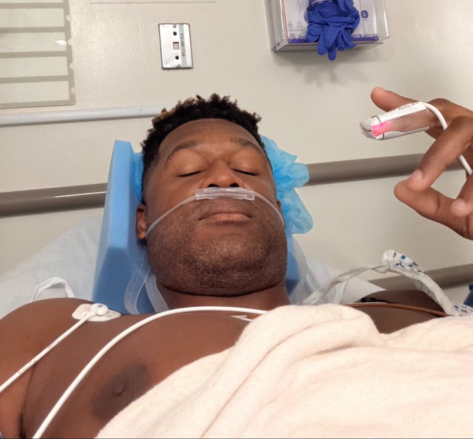 A man lies in a hospital bed with a nasal cannula and chest electrodes attached, holding up a pulse oximeter on his finger while giving a relaxed, eyes-closed smile—suggesting he's recovering well. The image humorously ties to the post's message that "the bull market may proceed," using his medical all-clear as a playful metaphor for the market's health despite any recent turbulence.