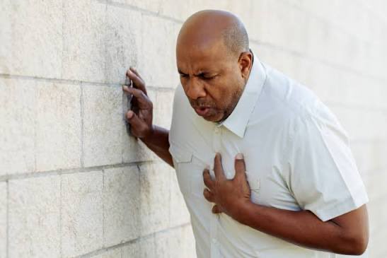 A bald man in a white short-sleeved shirt leaning against a stone wall, clutching his chest with a pained expression suggesting a heart attack.