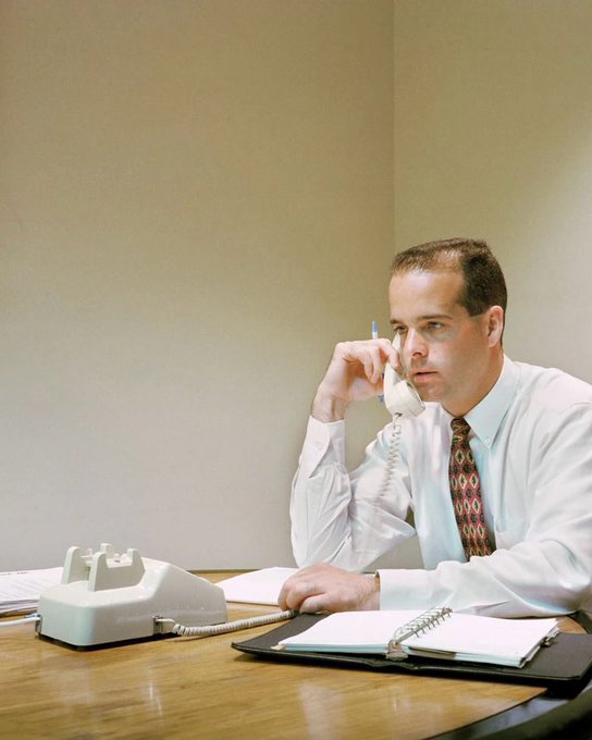 A photograph of a man in a white dress shirt and patterned tie sitting at a wooden desk, holding a pen near his face while using a white landline telephone.