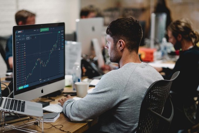 A man in a gray shirt sits at a desk in a busy office, looking at a stock chart on an iMac computer monitor.