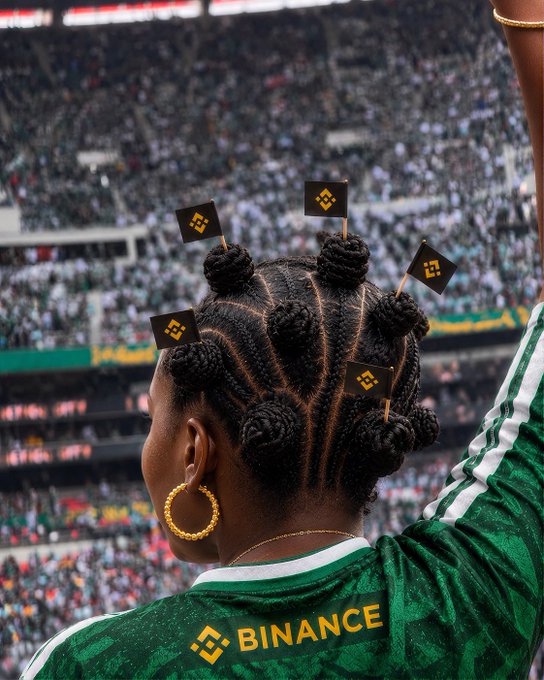 A person with braided hair styled in Bantu knots waves Binance-branded flags in a packed stadium, wearing a green jersey with "BINANCE" printed across the back—capturing the matchday energy from "the first whistle to the final celebration."
