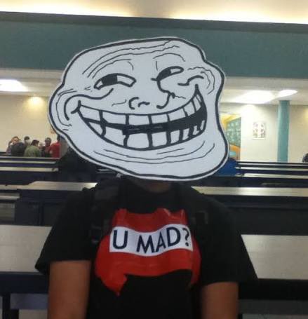 A person wearing a 'U MAD?' troll face t-shirt in what appears to be a cafeteria, with a smug troll face meme superimposed over their head.