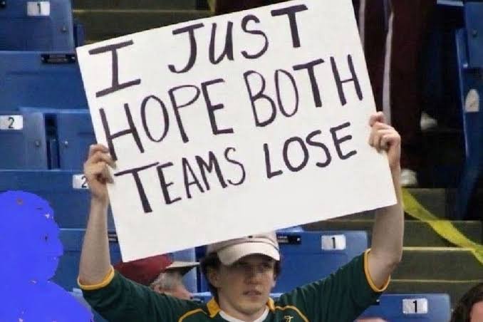 A sports fan wearing a green jersey holds up a handwritten sign that reads 'I JUST HOPE BOTH TEAMS LOSE' in empty stadium seats.