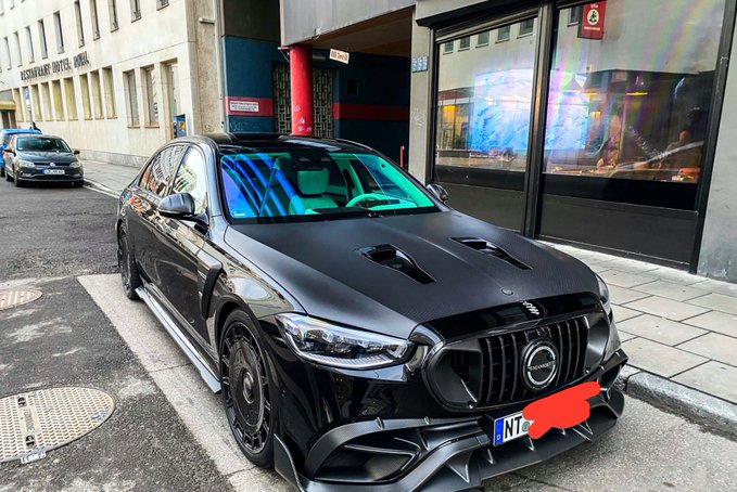 A sleek black Mercedes-AMG sedan is parked on a city street in front of an 'OKTAGON HOTEL' building.