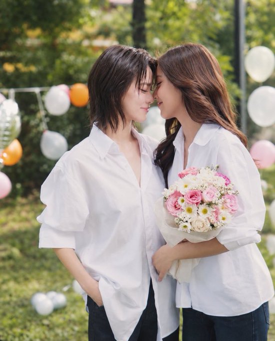 Two women in matching white button-up shirts stand closely together with their foreheads touching in an outdoor setting decorated with pastel balloons, one holding a bouquet of pink and white roses. The romantic pre-wedding styled scene aligns with the post's mention of "พรีเวดดิ้งแบบดื้อกระดุม" (pre-wedding with the top button undone) and "THE MOON ALWAYS BESIDE KEY #MoonshadowSeriesEP7," evoking an intimate, tender mood fitting for a BL series promotional shoot.