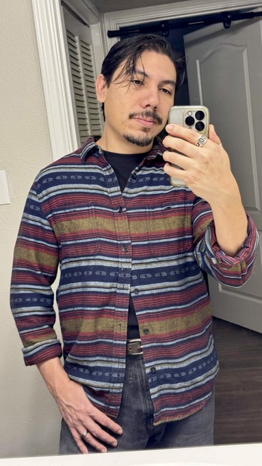 A man takes a mirror selfie wearing a colorful striped button-up shirt in shades of blue, burgundy, and olive green, while holding a phone displaying "00" on the screen. The casual photo appears to be a personal moment shared before his announced livestream appearance on the official OthersideMeta YouTube channel.