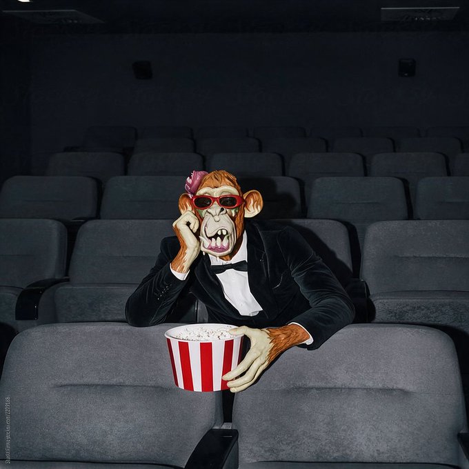 A digitally altered image shows a chimpanzee dressed in a black tuxedo with a red flower and red sunglasses, sitting alone in a dark movie theater and holding a red-and-white striped bucket of popcorn.