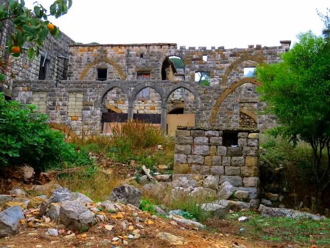 A dilapidated stone building with Gothic-style pointed arches stands in ruins, surrounded by overgrown vegetation and scattered rubble on the ground.