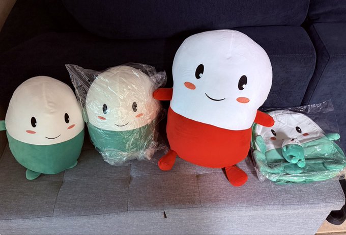 Four cute plush toys with smiling faces and small green or red bodies sit on a gray couch, two wrapped in plastic packaging.
