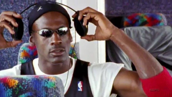 Michael Jordan in his Chicago Bulls uniform and Jordan-branded sunglasses holds a pair of headphones while sitting on a colorful patterned chair.