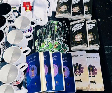 A collection of TFW (Tech For Women?) branded merchandise including white mugs, badges, lanyards, and notebooks spread on a dark surface.