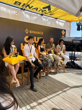 A panel discussion at a Binance event takes place on an outdoor stage under a yellow Binance-branded tent, with five panelists seated on stools holding microphones.