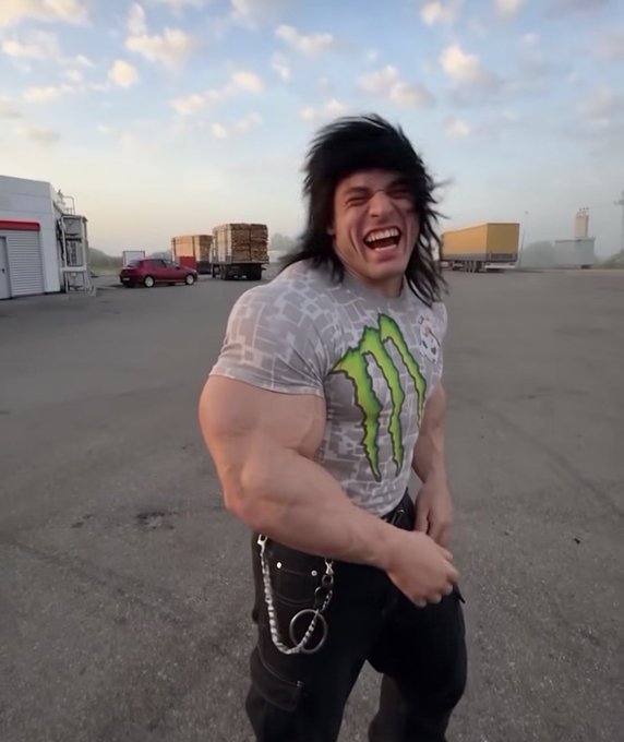 A muscular person in a black mullet wig and Monster Energy t-shirt laughs outdoors in what appears to be an industrial parking lot with semi-trucks in the background.