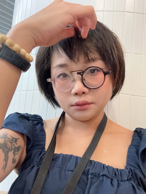 A selfie of a young woman with short black hair and large round glasses, wearing a dark blue off-shoulder top and showing a tattoo on her arm, posing with her hand near her forehead.