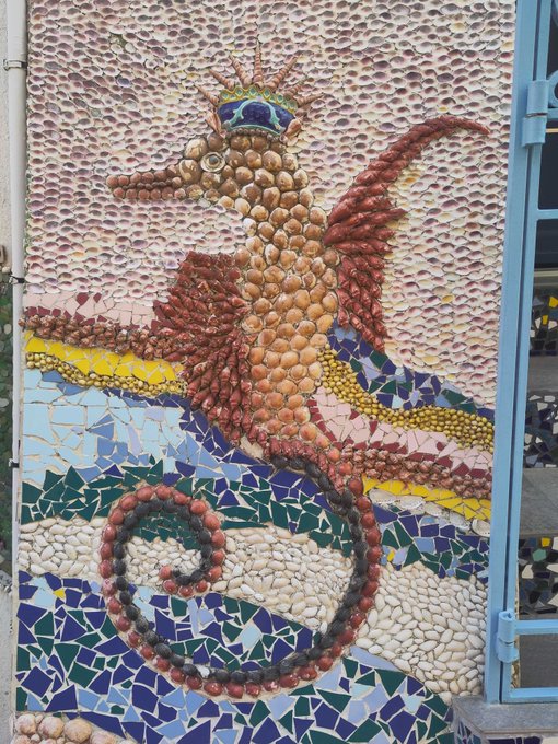 A colorful mosaic artwork depicting a seahorse made from various natural materials including seeds, beans, shells, and stones, set against a background of pink and blue tiled pieces.