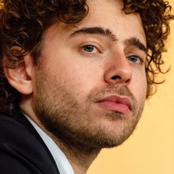 A close-up portrait of a young man with curly brown hair and light stubble, gazing thoughtfully at the camera against a soft yellow background.