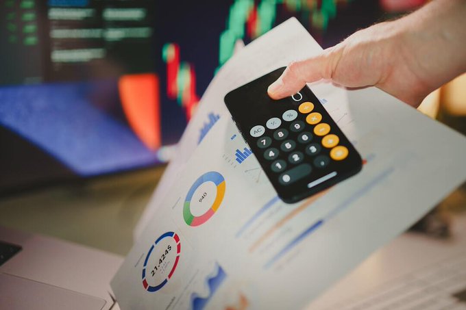 A close-up of a hand holding a calculator over printed financial charts and graphs, with a laptop and monitors displaying stock market data in the background.
