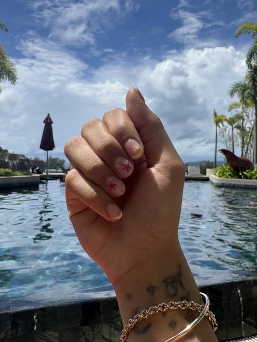 A hand displays pink manicured nails with small red and yellow star designs against a tropical poolside backdrop with palm trees and a partly cloudy sky. The image showcases the affordable Phuket nail art referenced in the caption, with gold bracelets and wrist tattoos adding to the vacation aesthetic.