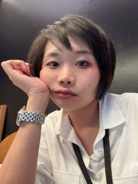 A woman with short dark hair wearing a white shirt and a watch poses for a selfie with her hand resting on her cheek.