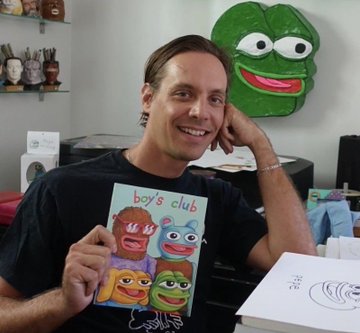 A smiling man holding a card with four colorful cartoon faces labeled 'boy's club', seated in front of a large green plush face on the wall.